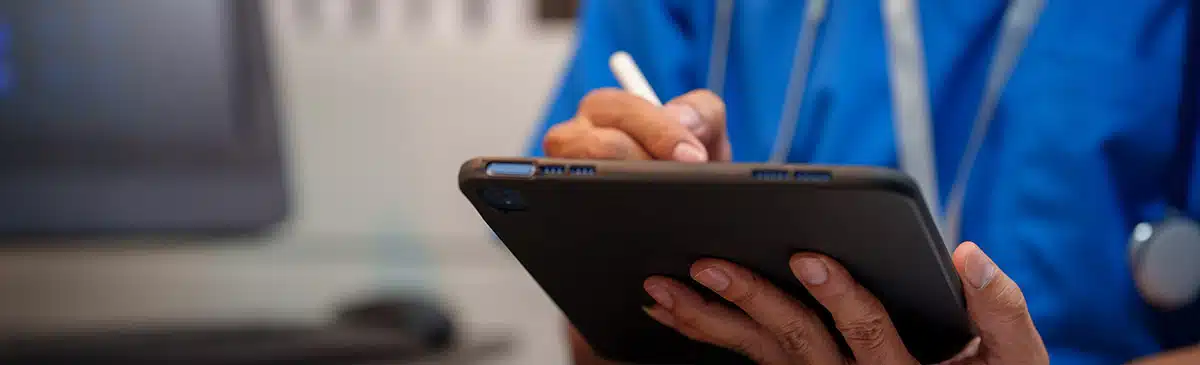 Close-up of heaalthcare worker in blue scrubs writing on a digital tablet with stylus at desk, stethoscope visible, computer monitor blurred in background, modern clinic office.